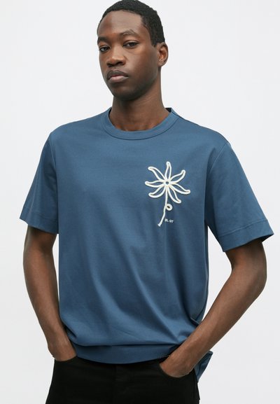 T-shirt de algodão azul com mangas curtas, apresentando um bordado floral branco no lado esquerdo do peito e um pequeno detalhe de texto por baixo.