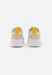Un paio di sneakers beige con suole bianche e linguette gialle brillanti sul tallone, mostrate dal retro su uno sfondo bianco.
