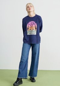 Sweatshirt bleu marine avec un imprimé graphique coloré, coupe décontractée, associé à un jean en denim à jambes larges et des bottes noires à lacets.