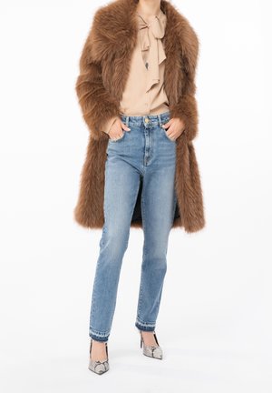 Abrigo de piel sintética marrón, blusa beige con detalle de lazo, jeans azul de cintura alta y tacones de serpiente con puntera afilada y tacones de aguja.