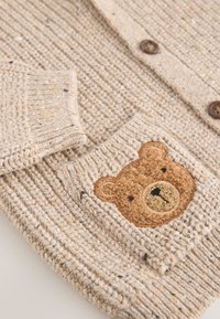 Cardigan en maille beige avec tissu texturé, présentant une broderie de visage d'ours sur la poche et des boutons en bois sur le devant.