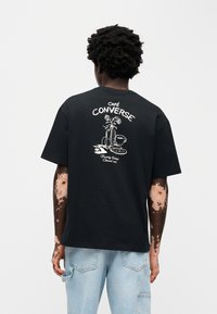 Μαύρο βαμβακερό t-shirt με λευκό γραφικό σχέδιο πίσω που απεικονίζει ένα βάζο με τριαντάφυλλα και μια κούπα καφέ. Χαλαρή εφαρμογή και κοντά μανίκια.