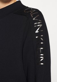 Černý pletený svetr s vysokým výstřihem. Na rameni se leskne reflektivní nápis "CALVIN KLEIN" v lesklém materiálu. Hladká textura.