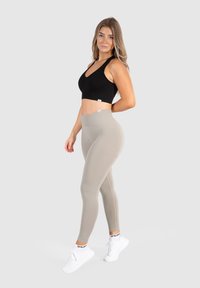 Schwarzer gerippter Sport-BH mit breiten Trägern, kombiniert mit hellbeigen, hoch taillierten Leggings. Das Model trägt weiße Sportschuhe mit Logo-Socken.