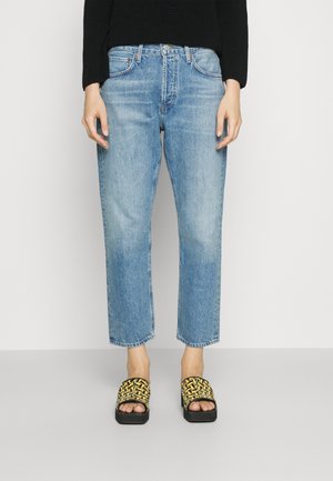 Osoba ubrana w jasnoniebieskie jeansy o prostych nogawkach, czarny sweter i czarne sandały na platformie z żółtymi plecionymi paskami, stojąca na białym tle.