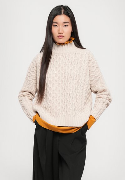 Cream knitted sweater with a cable pattern and high collar, layered over a mustard-colored top, paired with black wide-leg trousers.