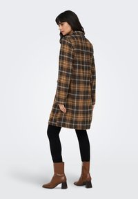 Palton maro în carouri, cu o siluetă lungă, având guler și buzunare. Purtat peste pantaloni negri și cizme maro cu toc. Textură fină a materialului.