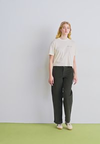 Witte katoenen t-shirt met een kleine print, gecombineerd met donkergroene broek. Model staat op een lichtgroene vloer, neutrale achtergrond.