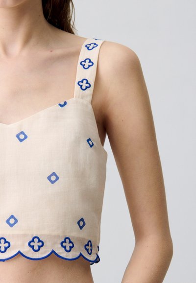 Haut court beige avec des motifs brodés bleus, présentant de larges bretelles ornées de motifs floraux et un ourlet festonné. Matière en lin.