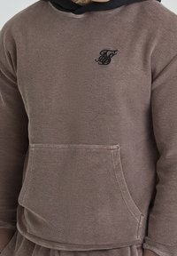 Sudadera marrón con acabado texturizado, capucha negra y bolsillo frontal. Presenta un logo bordado en negro en la zona del pecho.