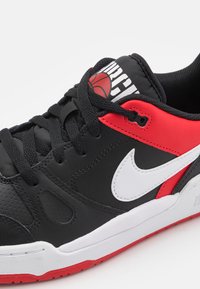 Sapatilha preta com detalhes vermelhos e o logo da Nike em branco. Apresenta couro texturizado, bico redondo e atacadores pretos rasos. Sola vermelha com padrão.