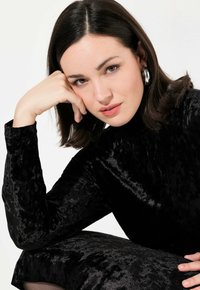 Top in velluto nero con collo alto e maniche lunghe, texture liscia e dettagli lucidi. Il modello ha capelli castani acconciati in modo morbido e trucco naturale.