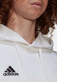 Haut à capuche blanc en tissu doux avec un cordon de serrage. Présente un logo brodé noir sur la poitrine et une texture lisse.