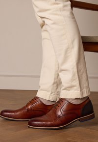 Braune Leder-DSchuh mit strukturiertem Finish, mit Schnürdesign, kontrastierendem dunklem Absatz und hellgrauen Socken, kombiniert mit cremefarbenen Hosen.