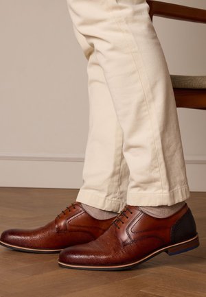 Braune Leder-DSchuh mit strukturiertem Finish, mit Schnürdesign, kontrastierendem dunklem Absatz und hellgrauen Socken, kombiniert mit cremefarbenen Hosen.