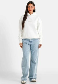 Felpa bianca con tasca frontale, caratterizzata da un piccolo logo rosa, abbinata a jeans azzurri larghi e scarpe bianche con accenti grigi.