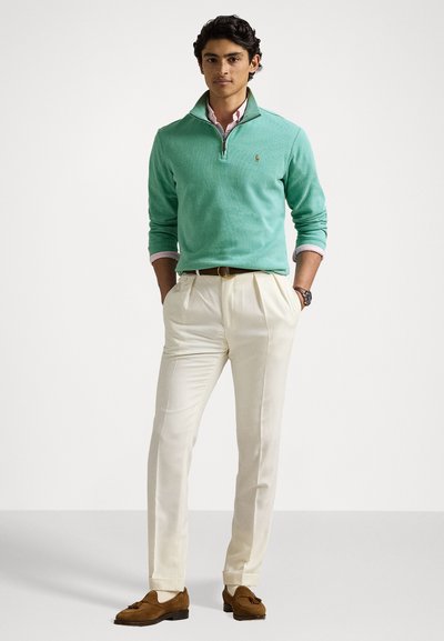 Μιντ πράσινο πουλόβερ με μισό φερμουάρ στον λαιμό, σε συνδυασμό με κρεμ παντελόνι και καφέ σουέτ παπούτσια. Γυρισμένα μανίκια, διακριτική λεπτομέρεια με λογότυπο.