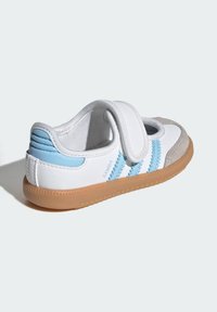 Biele športové topánky s modrými akcentmi, semišovou špičkou, suchým zipsom a textúrovanou hnedou gumovou podrážkou. Na boku má tri modré pruhy.