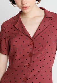 Vestido de color óxido con un cuello tipo camisa, mangas cortas, lunares negros y cinco botones rojos en el frente. Textura de tela suave.