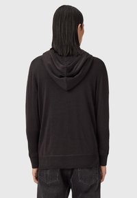 Person med axellångt mörkt hår iklädd en svart huvtröja och svarta jeans, med ryggen mot en enfärgad grå bakgrund.