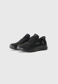 Chaussures de sport noires avec une tige en mesh, bout rond, semelle intercalaire texturée et semelle extérieure en caoutchouc plate. Comprend des lacets et un logo sur le côté et la languette.