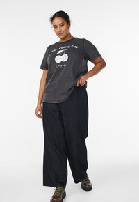 Frau mit lockigem Haar trägt ein schwarzes T-Shirt mit einer schwarzen-Kirsch-Grafik, weite schwarze Hosen und braune Stiefel und steht vor einem weißen Hintergrund.