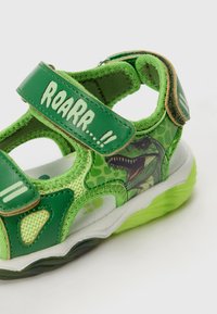 Sandale verte pour enfants avec un graphique de dinosaure, straps en Velcro, matériaux en maille et synthétiques, col rembourré et semelle texturée.