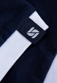 Marineblauer Baumwollstoff mit strukturierten Oberflächen, ausgestattet mit einem weißen Streifen und einem schwarzen Etikett mit weißem Branding. Deutliche Nähdetails sichtbar.