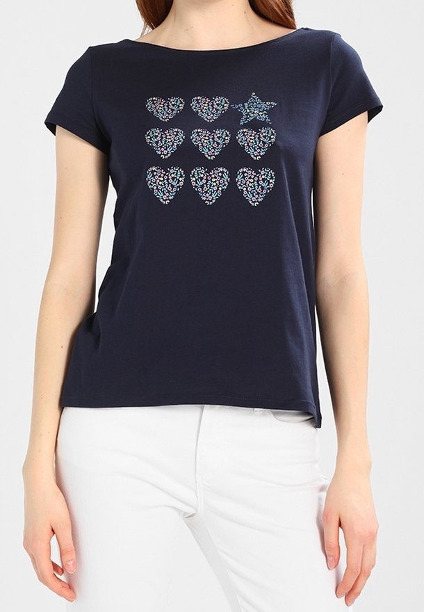 Femme portant un t-shirt bleu marine à manches courtes avec huit cœurs floraux et une étoile florale sur la poitrine, associé à un pantalon blanc.