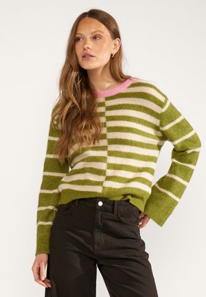 Frau mit langem, welligem Haar, die einen grün-weiß gestreiften Pullover mit pinkem Kragen und schwarze hoch taillierte Hose trägt, steht mit einer Hand auf der Hüfte.