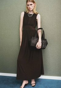 Brązowa bezrękawnikowa sukienka maxi z plisowanymi detalami, teksturowanym materiałem i zaznaczoną talią. Modelka trzyma małą czarną skórzaną torebkę na paskach.