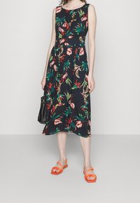 Ärmelloses schwarzes Kleid mit rotem und grünem Blumenmuster, tailliert und mit fließendem Rock. Kombiniert mit orangen flachen Sandalen und einer schwarzen Handtasche.