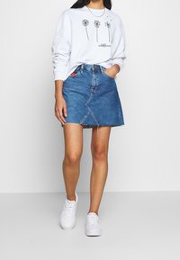 Gonna in denim con orlo grezzo, caratterizzata da un dettaglio di cucitura frontale e design a cinque tasche, abbinata a una felpa bianca con grafica e sneakers bianche.