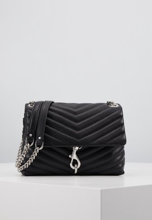 Schwarze gesteppte Lederhandtasche mit Chevron-Muster, silberner Kettenriemen und silberner Schnalle auf weißem Podest vor einfarbigem grauem Hintergrund.