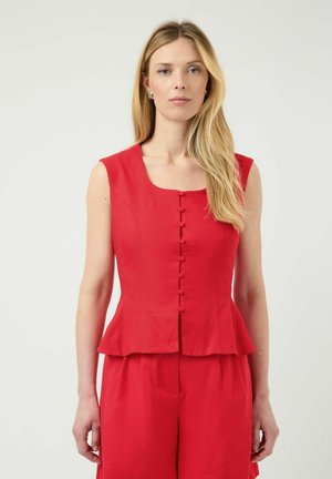 Blond kvinne som står, iført ermeløs rød knappet peplum-topp med matchende røde bukser foran ensfarget lys bakgrunn.