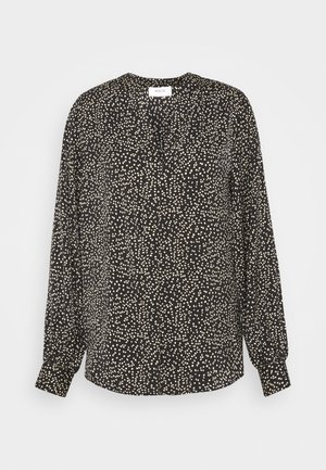 Blouse noire à manches longues avec des pois blancs irréguliers, col rond et poignets boutonnés, présentée sur un fond uni.