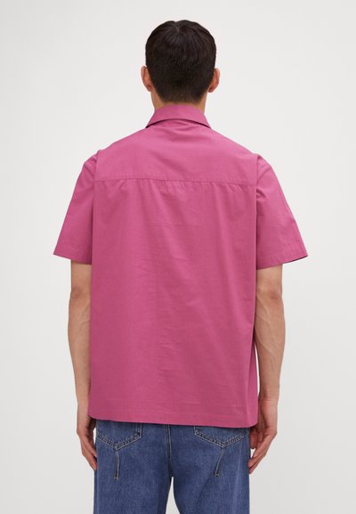 Camicia a maniche corte in tessuto rosa vivace, con colletto semplice, cucitura della spalla posteriore e vestibilità rilassata. Abbinata a jeans in denim blu.