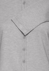 Cardigan gris clair avec une texture côtelée, cinq boutons sur le devant et un revers plié sur une manche.