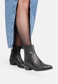 Botas de tobillo de cuero negro con un patrón de piel de serpiente texturizado, tacón bajo en bloque y paneles elásticos en los laterales, combinadas con una falda de mezclilla.