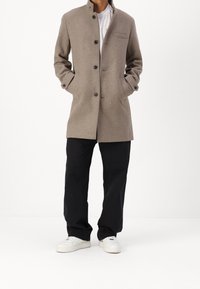 Abrigo de lana marrón claro con cuello alzado, cierre frontal de botones y un bolsillo en el pecho; llevado sobre pantalones negros y zapatillas blancas.