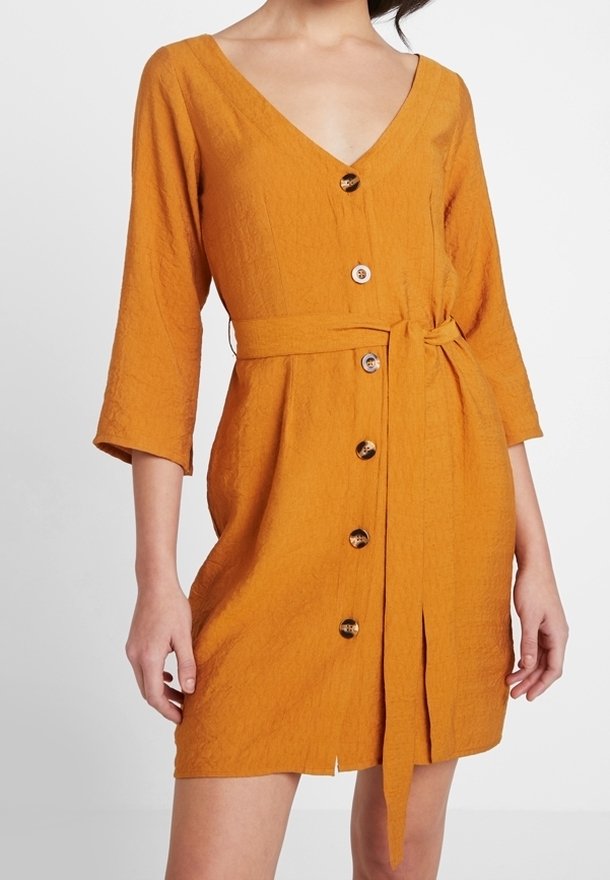 Ärm-lång klänning med V-ringning i strukturerad senapsfärgad tyg. Har en knappslå fram och ett matchande bälte i samma tyg.