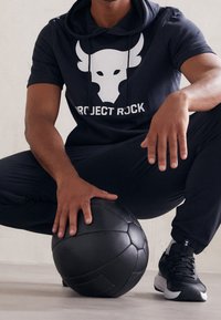 Musta lyhythihainen huppari, jossa valkoinen härkälogo ja "PROJECT ROCK" -teksti, yhdistetty mustiin urheiluhousuihin ja lenkkareihin, pitelee mustaa palloa.