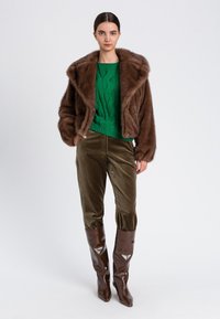 Giacca in pelliccia sintetica marrone, maglione a trecce verde, pantaloni in velluto a coste oliva e stivali marroni alti fino al ginocchio con punta a punta. Texture lisce, colori ricchi.