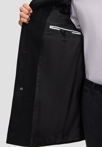 Interno del cappotto nero che mostra una fodera liscia con un dettaglio della tasca bianca a contrasto. Due bottoni visibili sulla parte esterna. Design e texture classici.