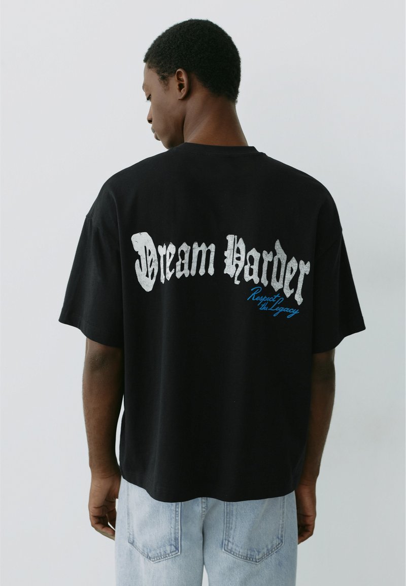 Homme portant un t-shirt noir oversize avec un texte blanc et bleu "Dream Harder Respect the Legacy" dans le dos, assorti à un jean bleu clair.