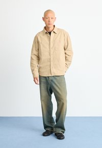Camisa de algodón beige con botones en la parte delantera y dos bolsillos en el pecho, combinada con unos pantalones vaqueros azules holgados y zapatos negros sobre un suelo azul.