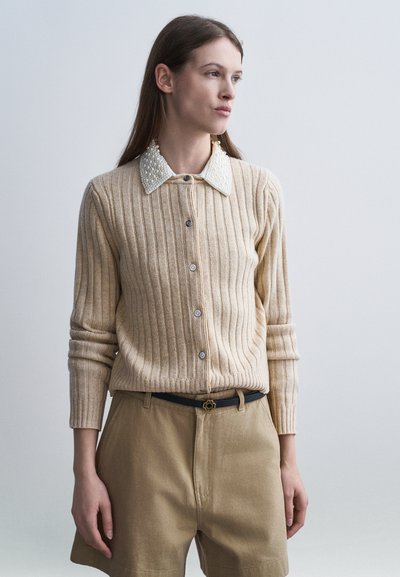 Mujer con un cárdigan beige acanalado con cuello de perlas y pantalones cortos beige de cintura alta, de pie frente a un fondo claro y liso.
