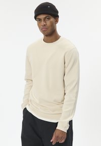 Béžová mikina s dlhými rukávmi, okrúhlym výstrihom a rebrovanými manžetami, doplnená tmavou pletenou čiapkou. Jednoduchá, hladká textúra.