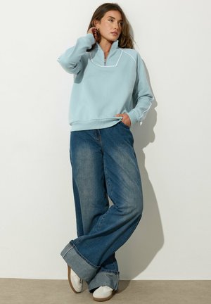 Light blue sweatshirt with a half-zip collar, paired with loose-fitting, faded blue jeans rolled at the cuffs. Sneakers complete the look.