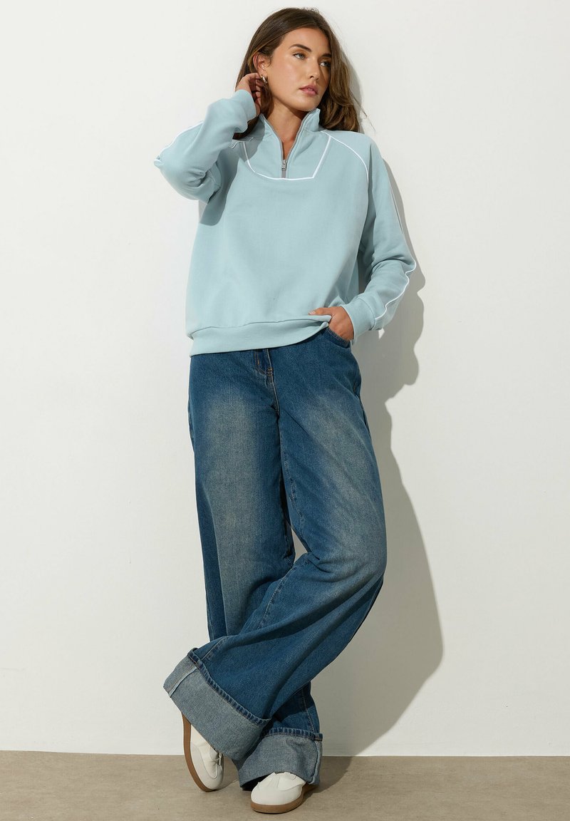 Felpa azzurra chiara con colletto a metà zip, abbinata a jeans blu sbiaditi e a taglio largo arrotolati in fondo. Le sneaker completano il look.